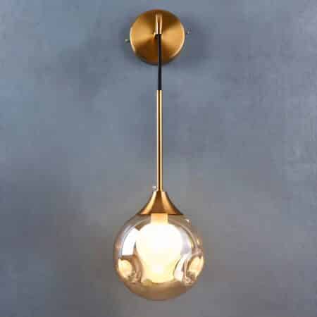 Gold-plated metal wall sconce with suspended glass ball. The ball is not smooth but has 2 recesses on each side, the bulb is lit, the wall light is fixed to a grey wall.