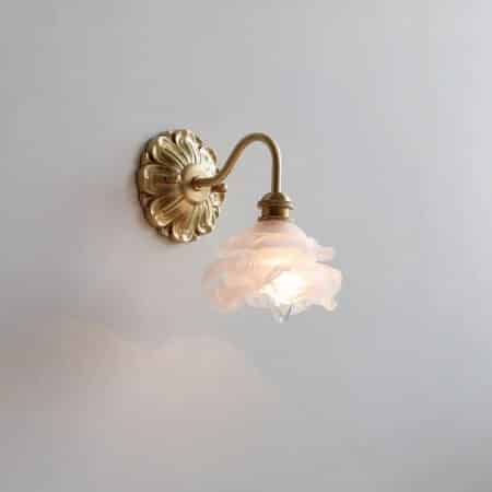 Vintage wall sconce with gilded metal frame holding a suspended glass rose. The wall light is lit and mounted on a gray wall.
