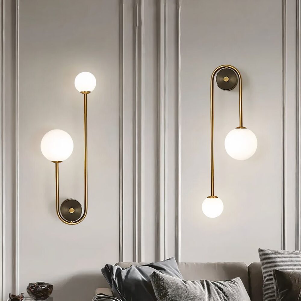 Grey wall on which 2 wall sconces are fixed, round gilded support with a gilded rod running left and right, one shorter than the other. On the smaller one, a large luminous ball, on the longer one, a small luminous ball. The sconce on the left is fixed ball upwards, the sconce on the right ball downwards. Beneath the sconces is a grey sofa with matching cushions.