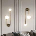 Grey wall on which 2 wall sconces are fixed, round gilded support with a gilded rod running left and right, one shorter than the other. On the smaller one, a large luminous ball, on the longer one, a small luminous ball. The sconce on the left is fixed ball upwards, the sconce on the right ball downwards. Beneath the sconces is a grey sofa with matching cushions.