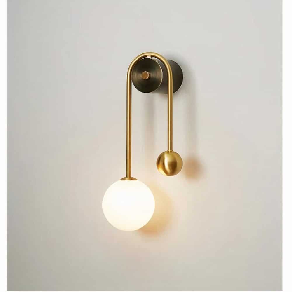 Wall sconce mounted on a white wall, round gilded support, gilded stem on either side of the support with a luminous ball on the left and a smaller gilded ball on the right.