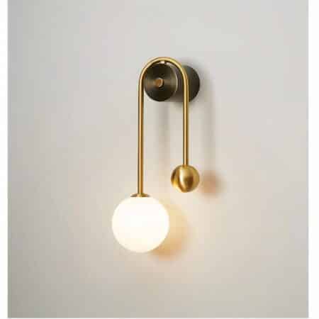 Wall sconce mounted on a white wall, round gilded support, gilded stem on either side of the support with a luminous ball on the left and a smaller gilded ball on the right.