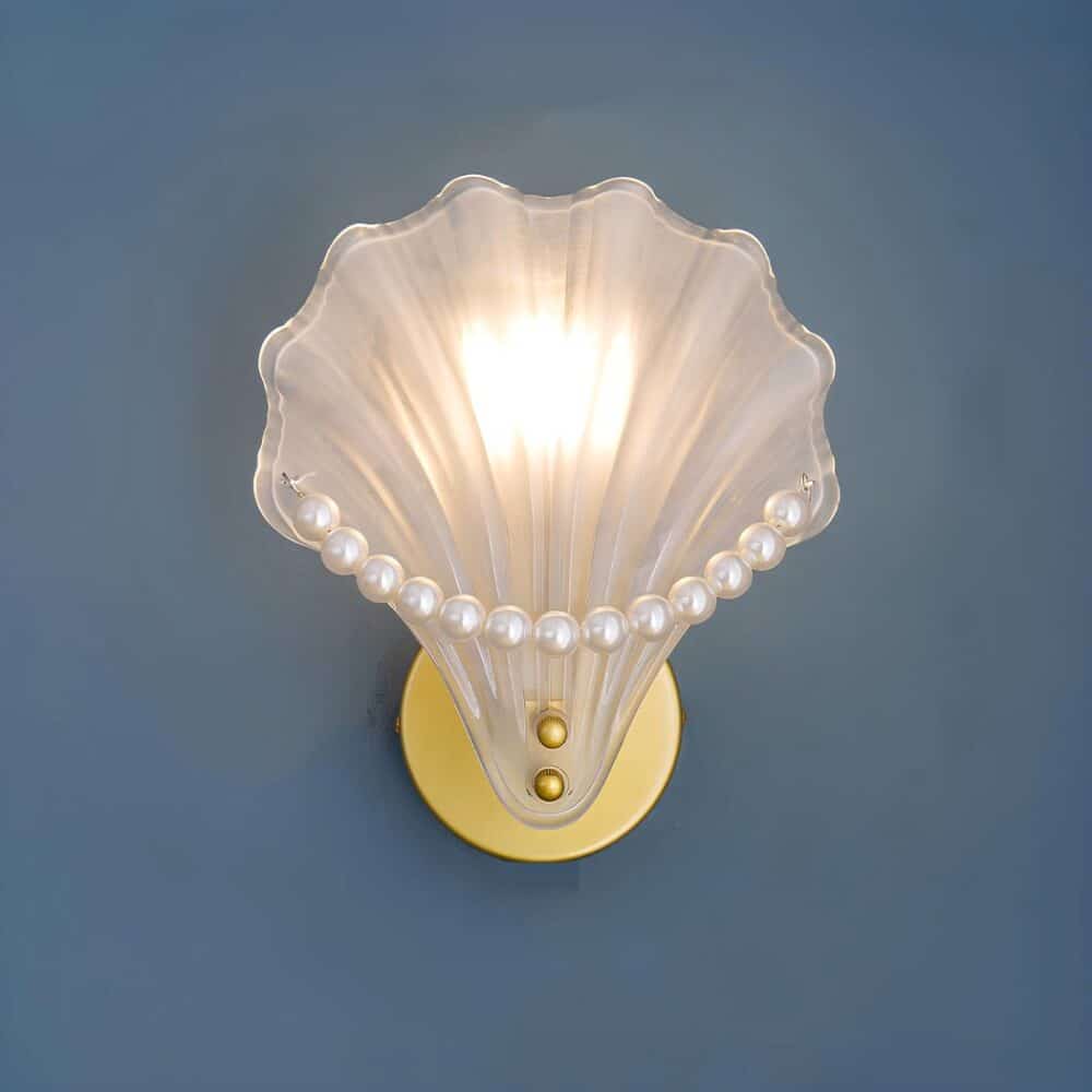 Wall sconce in the shape of a shell in polished glass with white pearls on the edge of the shell. The round gold bracket is mounted on a blue wall. The bulb is lit.