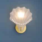 Wall sconce in the shape of a shell in polished glass with white pearls on the edge of the shell. The round gold bracket is mounted on a blue wall. The bulb is lit.