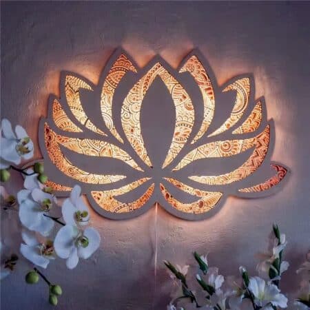 Mandala-style lotus flower wall sconce lit up, allowing light to pass through its openwork pattern. Geranium-style flowers are visible in the bottom corners of the picture.