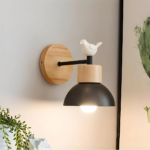 Modern black wall lamp in wood with a bird that is lit, next to a picture and a fake plant