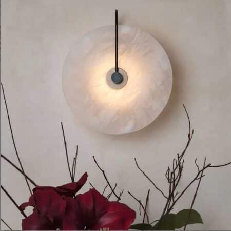 Wall sconce with marble wheel and lit black stem, mounted on a white wall. Dark pink branches and flowers can be seen under the sconce.