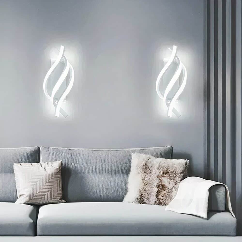 2 white spiral 3-strand wall sconces lit in cold light. Mounted on a grey wall, dark grey curtain on the right. A gray sofa under the sconces with 2 cushions: one white with gold pattern and one with fur, fabric on the sofa armrest.
