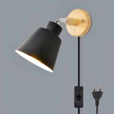 Wall light mounted on a gray wall, its support is made of wood, its shade is made of black aluminum, it has a connecting cable with a switch and its EU plug is visible. The bulb is lit.