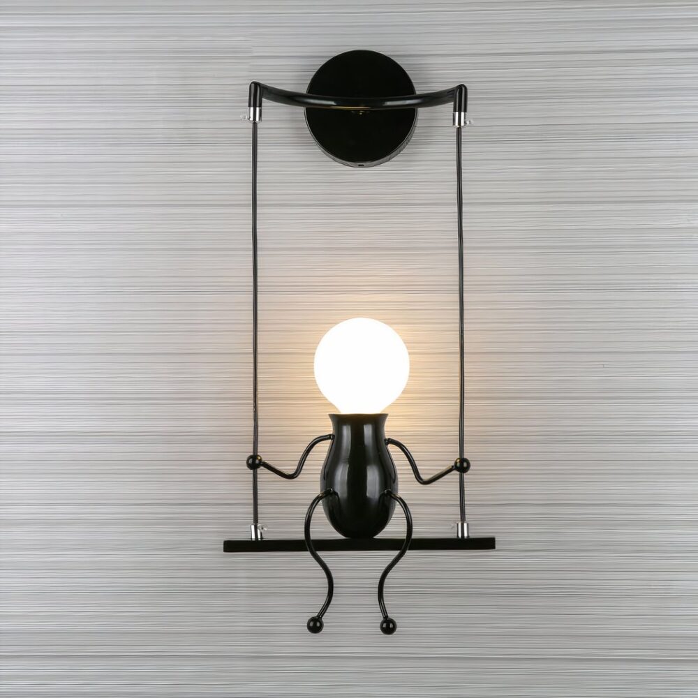 Wall light in the shape of a swing with a man sitting on it. His head is the light bulb. It is mounted on a gray wall