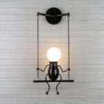 Wall light in the shape of a swing with a man sitting on it. His head is the light bulb. It is mounted on a gray wall