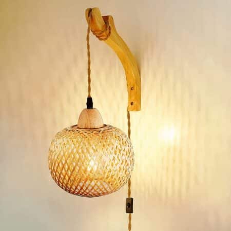 Suspended wall light with wooden bracket fixed to a white wall and openwork bamboo oval connected by a rope which can also be seen under the bracket with a switch. Bulb on.