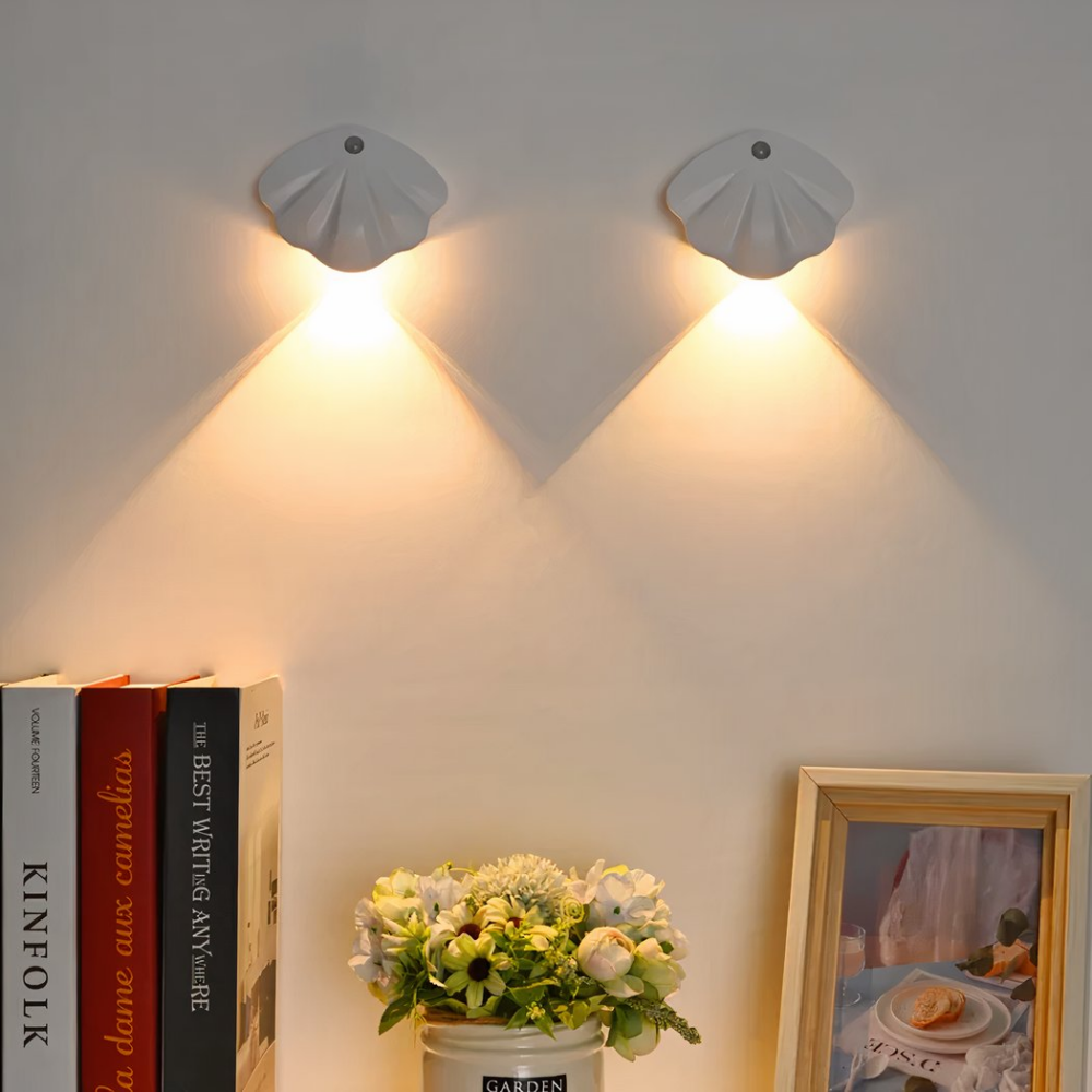 2 small wall sconces in the shape of a white seashell, illuminated towards the white, where there are books on the left, a small green plant in the center and a picture frame on the right.