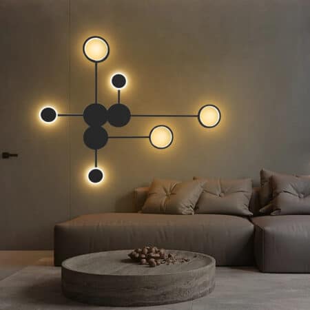 Black wall light with circles linked by lines. 3 black circles in the center then 6 circles around luminous. Mounted on a brown wall above a brown sofa with matching cushions. Brown round coffee table and beige carpet underneath.