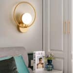 Circle-shaped gold wall light with gold spotlight on lower part. To the right is a 2-door cupboard with white mouldings, below the wall light is a bedside table with a book and a small plant, and to the left is a gray bed with blue and black cushions.