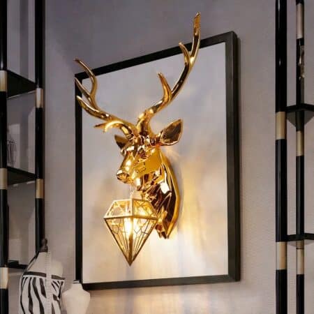 Large wall sconce in the shape of a golden stag bust holding in its mouth a diamond-shaped suspension in which the light bulb is lit. Wall-mounted on a black frame with white background. Black and gold shelves on the left and right.