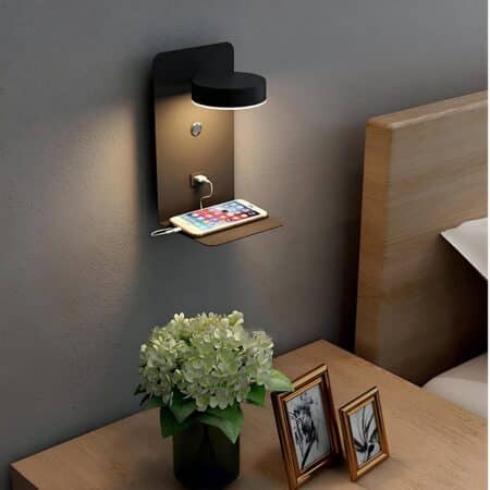 Rectangular black wall light consisting of a round adjustable spotlight, a switch, a USB socket and a shelf for placing objects, all fixed to a gray wall. Below, a bedside table with 2 small frames and a green plant, and to the right a bed with a brown headboard. The spotlight is on.