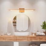 Long wooden wall lamp lit above a round mirror. The mirror is above a white washbasin on a wooden top with bottles to the left and right of the washbasin, beige towels to the right and a green plant.