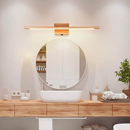 Long wooden wall lamp lit above a round mirror. The mirror is above a white washbasin on a wooden top with bottles to the left and right of the washbasin, beige towels to the right and a green plant.