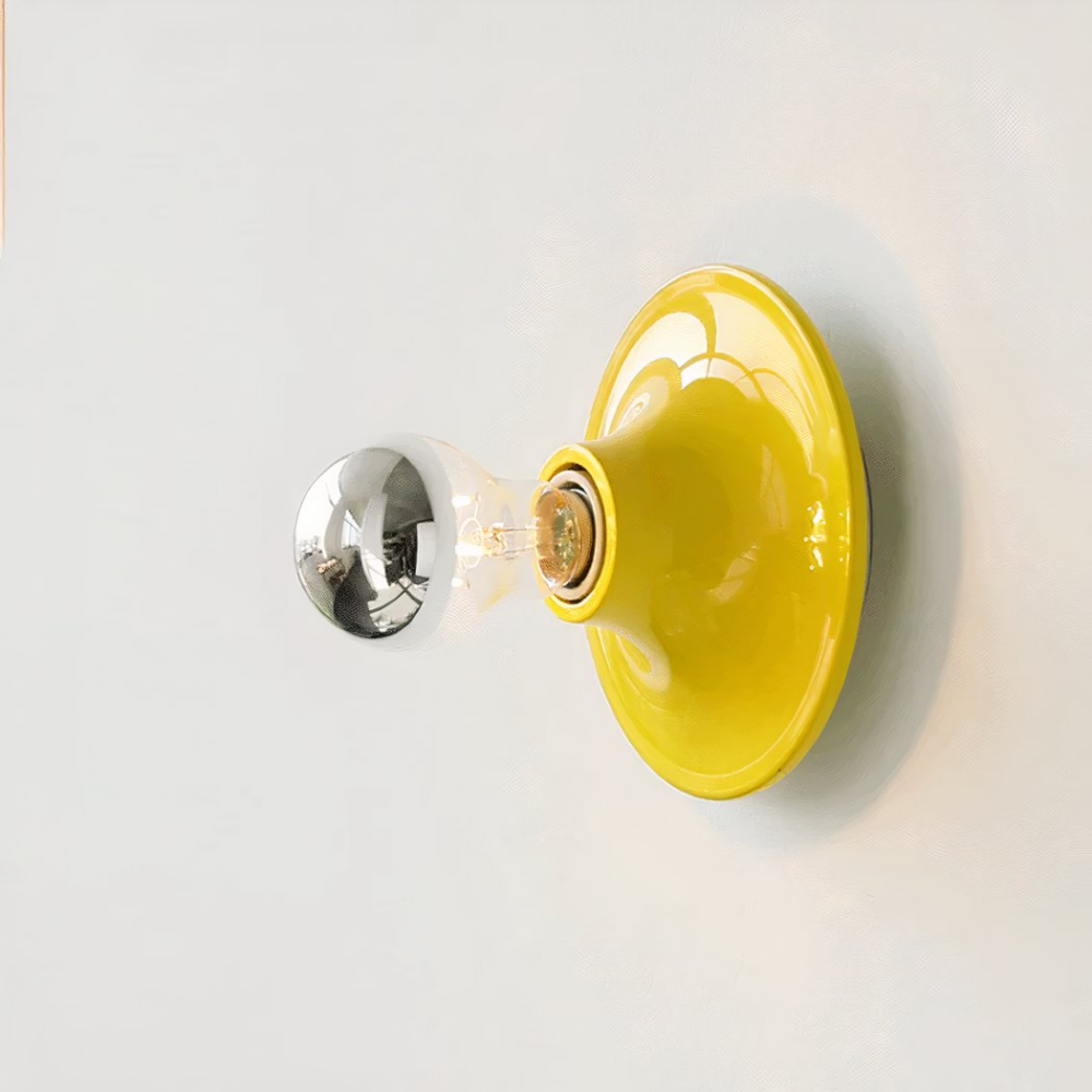 Round, yellow wall light resembling a small plate with a socket in the center. A bulb with a silver top is screwed in. The sconce is mounted on a white wall.