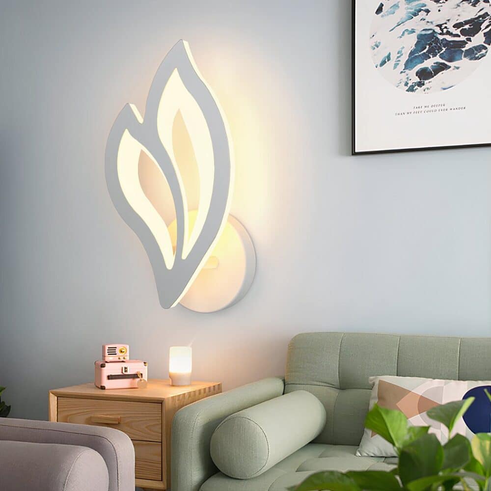 Petal-shaped wall light in white acrylic, diffusing a warm light. It is mounted on a gray wall above a small bedside table with a pink alarm clock. To the right, a green sofa with a white and pink cushion. Above the sofa, an abstract frame. In the lower left-hand corner, a corner of a grey armchair.
