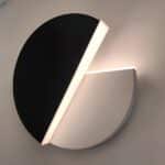 Wall light in 2 half-moon parts, one of which is black and rotatable. The other fixed part is white. Light is diffused by the edges of the half-moons. The rotating part is open at approximately 45 degrees.