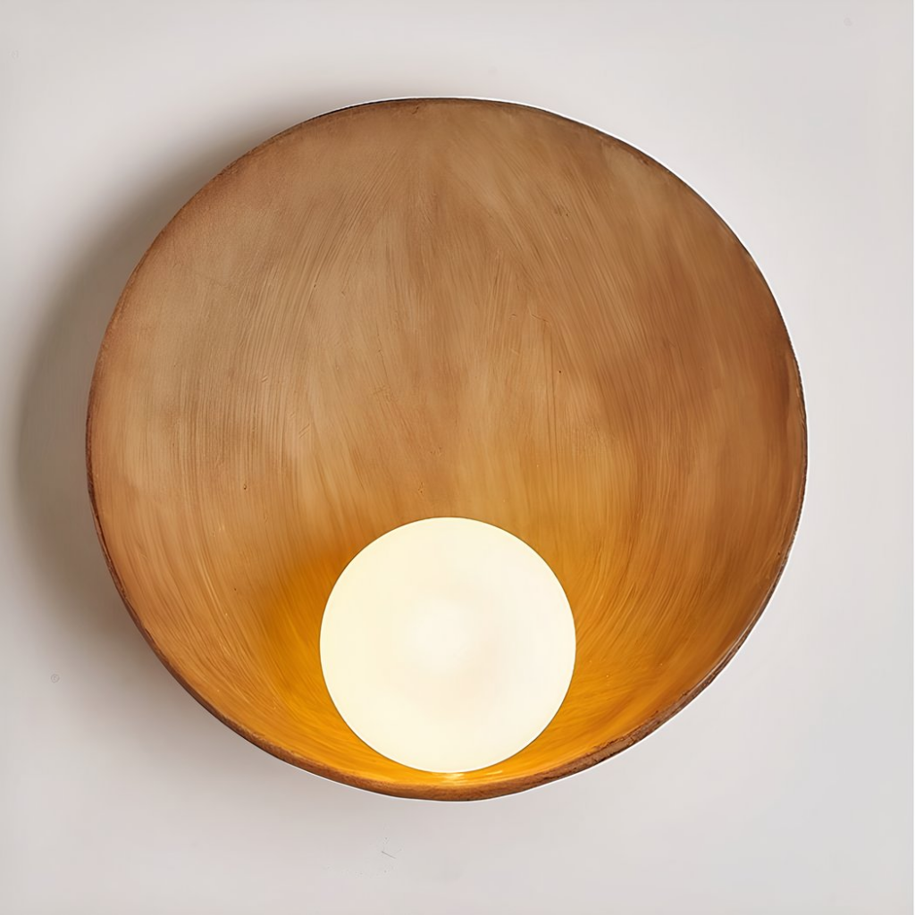 Round wall lamp in imitation wood resin, giving the impression of a shell with a luminous ball on the bottom. Mounted on a white wall.
