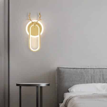 Wall sconce with luminous gold circle and luminous gold oval in front and deer head at top in gold. Mounted on a white wall. Grey sofa and black table.