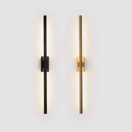 2 tube-shaped wall sconces mounted vertically on a white wall. The sconce on the left is black, the sconce on the right is gold. Light is diffused only through the part of the tube facing the wall.