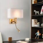 Wall sconce with rectangular gold base and rectangular upward bent stem. White fabric shade above. The bulb is lit. On the bracket, two black switches and a gold flexible reading lamp. The wall sconce is mounted above a brown desk and to the left of a brown bookcase containing books and figurines.
