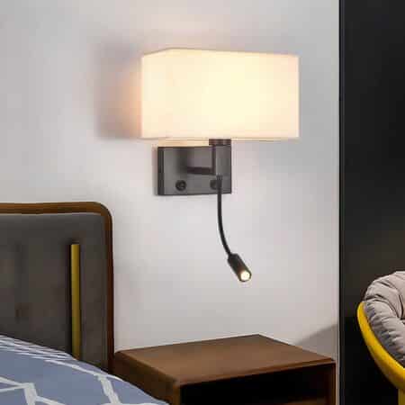 Vintage wall lamp with black rectangular base, black angled stem and beige fabric shade. Two switches and a lit flexible reading lamp are mounted on the bracket. The sconce is mounted above a brown bedside table. To the left, a brown bed and to the right, a round yellow armchair with a grey cushion.