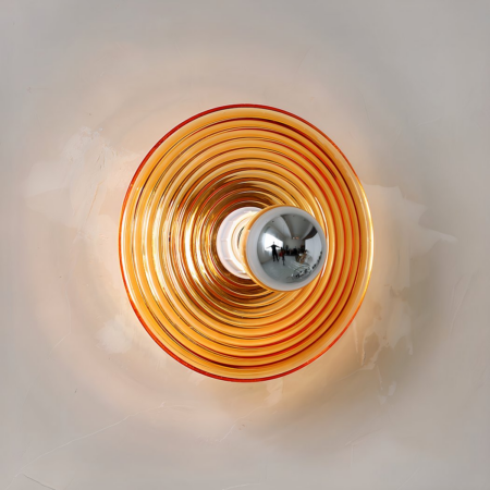 Retro-style wall light with orange glass circle with grooves in circles from smallest to largest towards the outside. The bulb is fixed in the center with its silver-colored head. Mounted on a gray wall.