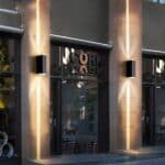 Storefront with display windows framed by concrete pillars on each of which a wall sconce is mounted. The wall light is rectangular and black in color, diffusing a beam of warm light above and below.