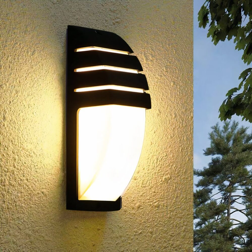 Half-moon shaped wall light with black structure, one part covers the top of the half-moon with spaces allowing light to pass through. Mounted on a beige wall. A tree and blue sky can be seen on the right.