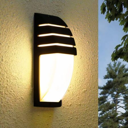 Half-moon shaped wall light with black structure, one part covers the top of the half-moon with spaces allowing light to pass through. Mounted on a beige wall. A tree and blue sky can be seen on the right.
