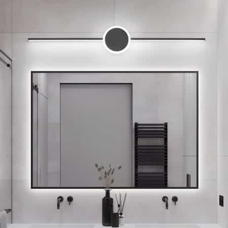 Wall light in the form of a long black tube with a black circle in the center. Light is diffused through the back of the tube and around the circle. The wall light is mounted in a bathroom above a mirror, on a white tiled wall with a washbasin below. In the mirror, the reflection of a door and a black towel rail.