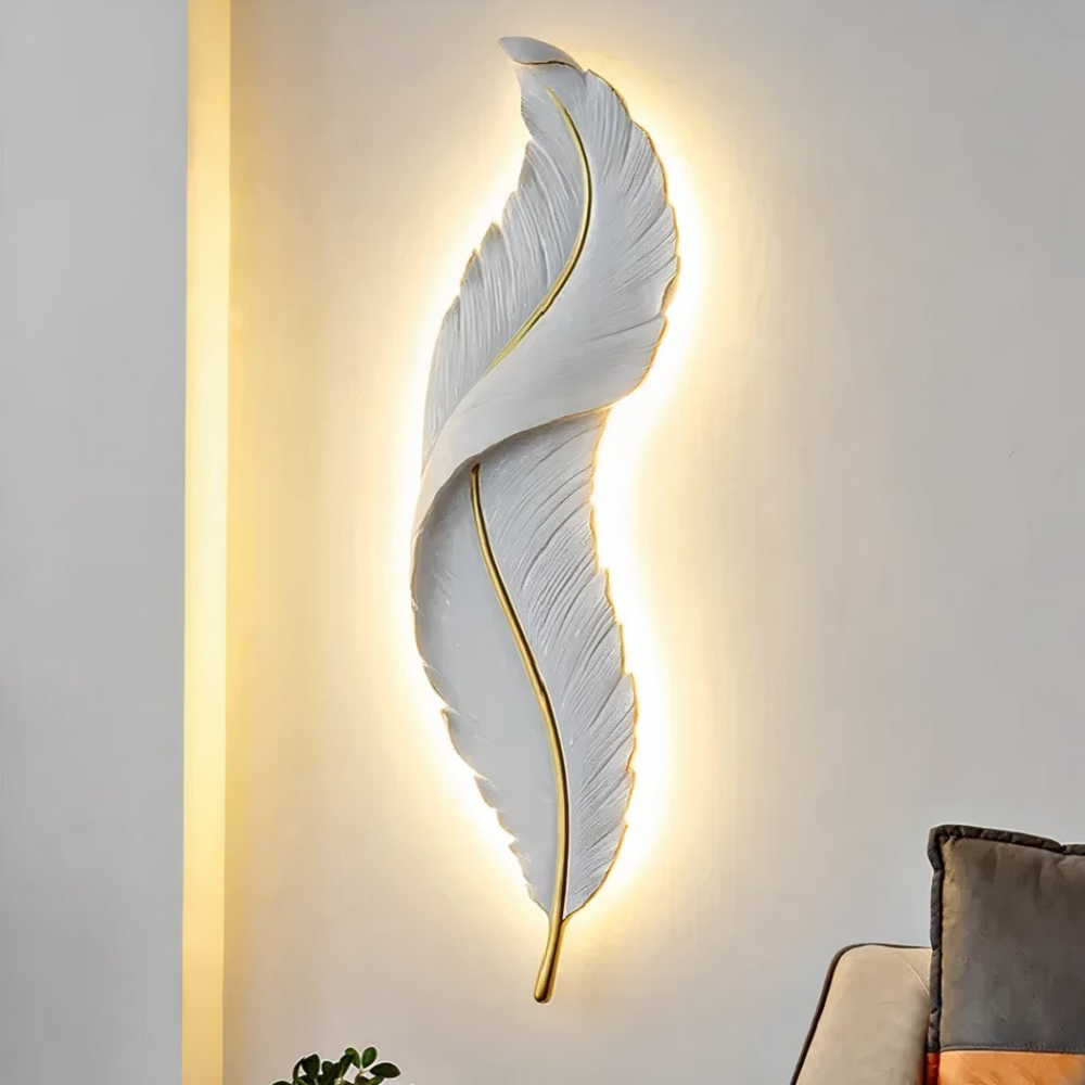 White feather wall light with gold center and tail, diffusing a warm light at the back. Mounted on a white wall with a wall corner on the left and a brown sofa on the bottom right with brown cushion.