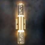 Wall sconce in the shape of a bubble tube with gold support and gold part in the center of the tube. Fixed on a black wall. The wall light is switched on and emits a warm light through the tube.