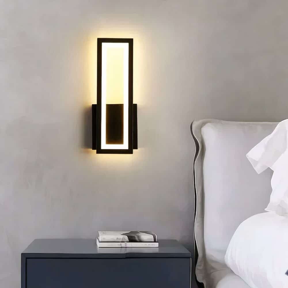 Black wall light, rectangular in height. Mounted on a grey wall, lit above a black bedside table, with a book on top and a white bed to the right.