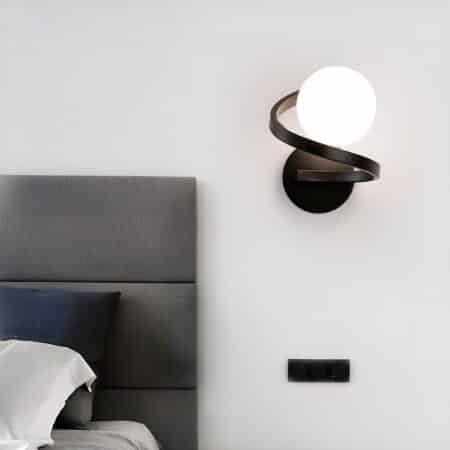 Wall light with black spiral and ball light at top. Mounted to the right of a bed with gray headboard on a light gray wall.