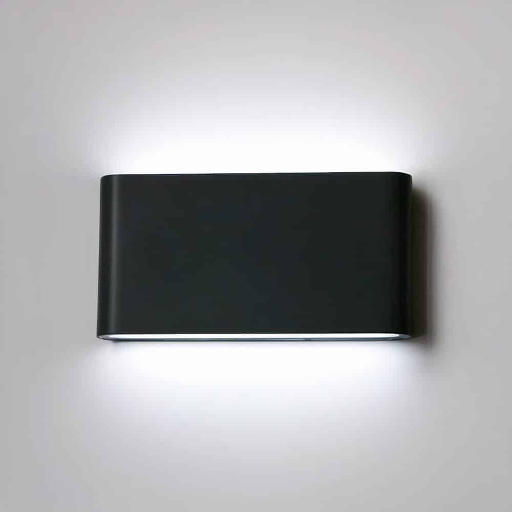 Black wall sconce with rounded rectangular shape, light is diffused from the top and bottom of the sconce. It is switched on and mounted on a white wall.