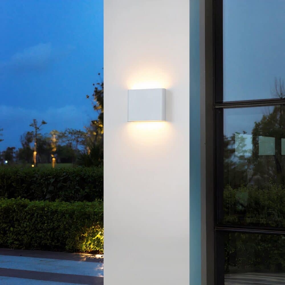 White wall light with rounded rectangular shape mounted on a white exterior wall. A window on the right with black frame. Bushes in background on the left.