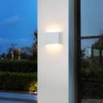 White wall light with rounded rectangular shape mounted on a white exterior wall. A window on the right with black frame. Bushes in background on the left.