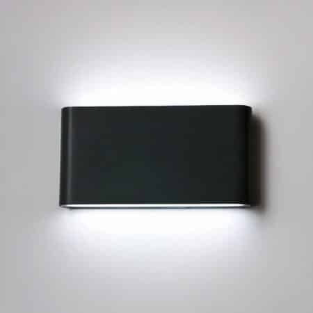 Black wall sconce with rounded rectangular shape, light is diffused from the top and bottom of the sconce. It is switched on and mounted on a white wall.