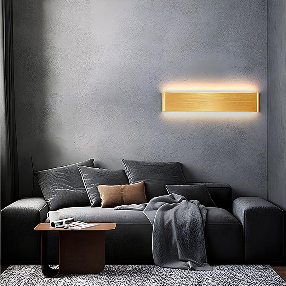 Rectangular gold wall light mounted on a dark gray wall. It is lit and above a gray sofa with gray cushions, a small brown table in front of the sofa and a beige rug.