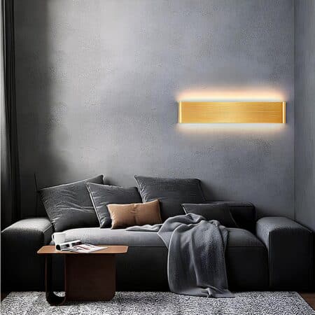 Rectangular gold wall light mounted on a dark gray wall. It is lit and above a gray sofa with gray cushions, a small brown table in front of the sofa and a beige rug.
