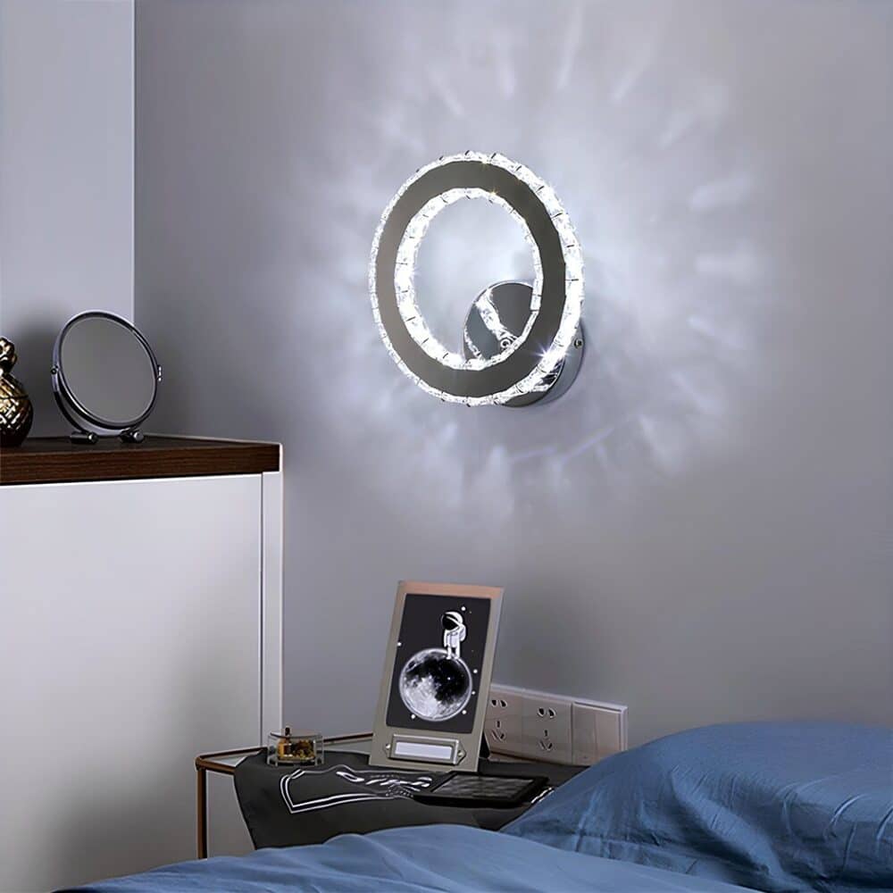 Circle-shaped wall light with inner and outer contours imitating diamonds. The light is diffused by these diamonds with a cool glow. The wall light is fixed above a bedside table with gray fabric and a frame with electrical sockets behind it. To the right, a bed with blue bedding. To the left, a white wall unit with a round mirror.