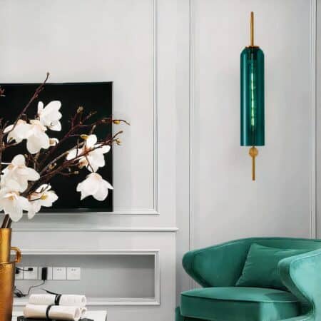Art Deco-style glass tube wall light lit and hung on a white living room wall, above a forest green armchair.