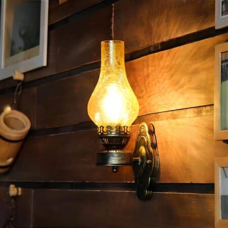 Vintage wall light in the shape of an oil lamp in cracked yellow glass, bulb on. It is mounted on a horizontal wooden slat wall, with a white frame on the left-hand corner and a bamboo-style tube underneath.