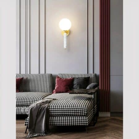 Ball wall lamp in colored marble and gilded brass in its white version, lit and hung on the gray wall of a living room, above a houndstooth print sofa.